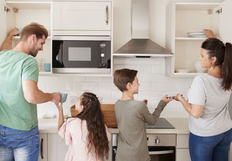 En una cocina, la figura de un hombre ayudando a una niña, y la figura de una mujer ayudando a un niño. Se interpreta como una familia colaborando en conjunto. Sonríen.