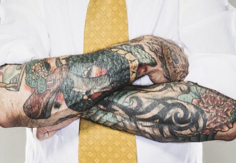 Brazos cruzados, tapados casi en su totalidad por tatuajes; la persona viste camisa y corbata.