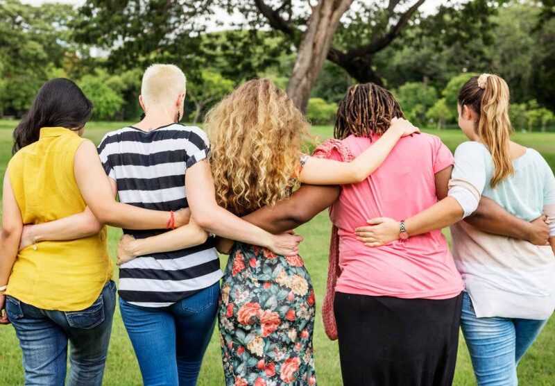 Cinco personas diferentes, de espalda tomándose de los brazos abrazándose en un escenario de naturaleza, parque verde.